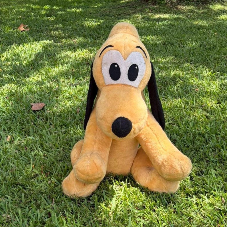 May include: A plush toy of Pluto, a yellow dog with black ears and a black nose, sits on green grass. The toy has large white eyes with black pupils and is a popular character from Disney.