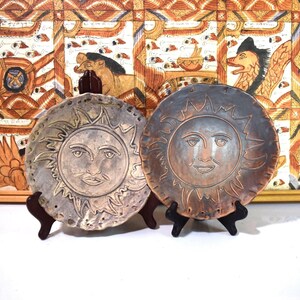 May include: Two ceramic plates with sun faces, one silver and one brown, displayed on small wooden stands. The suns have detailed facial features and are surrounded by radiating lines. The background features a colorful, patterned artwork.