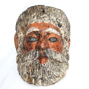 May include: A weathered, hand-carved mask depicting a bearded face with a stern expression. The mask features a reddish-brown face, white hair and beard, and dark, hollowed-out eyes. The mask is likely made of wood or a similar material.