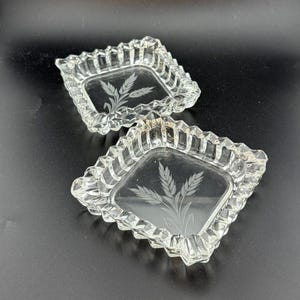 May include: Two clear glass ashtrays with a square shape and a decorative, scalloped edge. Each ashtray features an etched wheat design in the center. The ashtrays are displayed on a black surface.