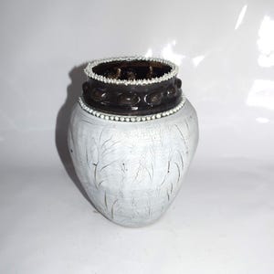 May include: A white ceramic vase with a cracked, textured surface. The vase has a dark brown band with decorative circles and a rim adorned with small white beads. The top rim is also dark brown with a textured interior.