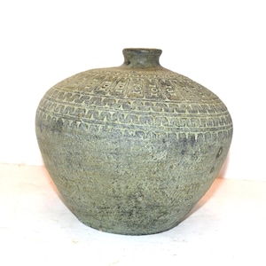 May include: A round, wide-bodied ceramic vase with a narrow neck and a textured, weathered green and grey finish. The upper portion of the vase features horizontal bands of repeating geometric patterns, including small rectangles and zigzag lines.