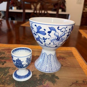 May include: Two white and blue porcelain egg cups. The larger cup has a floral design, while the smaller cup features a pagoda scene. Both have a pedestal base and are set on a decorative mat.