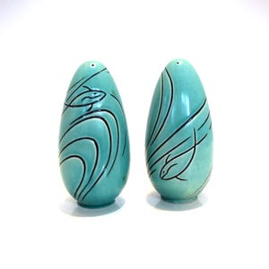 May include: A pair of turquoise ceramic salt and pepper shakers. Each shaker is egg-shaped with a black fish and wave design. The shakers have a glossy finish and small holes at the top for dispensing.