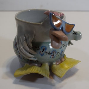 May include: A ceramic chicken mug with a floral design and yellow feet. The chicken is wearing sunglasses and has a pink flower on its head.