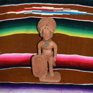 May include: A terracotta-colored clay figurine of a standing figure wearing a large, textured headdress and a circular pendant. The figure holds a rectangular shield with a carved central design in one hand and stands on a flat base.