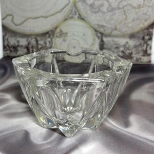 May include: A clear, faceted glass ashtray with a geometric design. The ashtray has a square shape with angled sides and a central depression. The glass reflects light, creating a sparkling effect. The background features a vintage map.