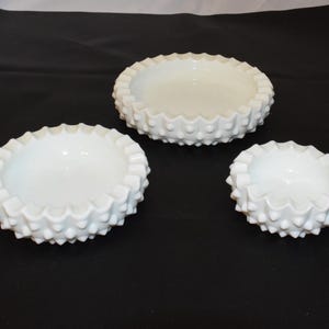 May include: Three white, textured, decorative bowls of varying sizes. Each bowl has a scalloped edge and a pattern of small, raised bumps around the exterior. The bowls are arranged on a dark surface.
