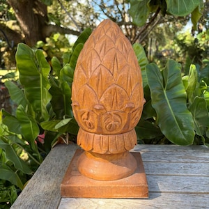 May include: A carved wooden decorative pineapple sculpture. The sculpture is a warm brown color and features detailed carvings. It is mounted on a square base and sits on a wooden table. The background includes green foliage.
