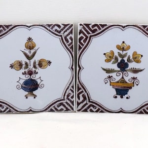 May include: Two square ceramic tiles with floral designs. Each tile features a white center with a stylized flower arrangement in yellow, blue, and brown. The edges are framed with a dark brown geometric pattern.