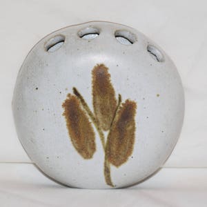 May include: White ceramic vase with a brown and green floral design. The vase has five holes around the top for holding flowers.