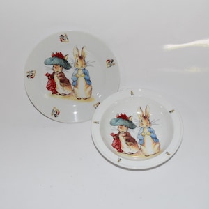 May include: Two white ceramic plates with a Peter Rabbit design. The larger plate features two rabbits wearing blue jackets and red hats. The smaller plate is a bowl with the same rabbits.
