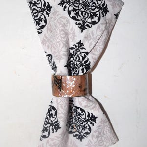 May include: A folded white and gray patterned napkin with black floral designs is held by a copper-colored napkin ring. The napkin has a gray border.