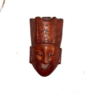 May include: A carved wooden mask with a smiling face and a tall, ornate headdress. The mask is a rich, reddish-brown color and features detailed carvings, including a symbol on the headdress. The face has a serene expression.