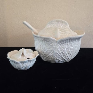 May include: A white ceramic cabbage-shaped serving bowl with a matching smaller bowl and a ladle. The bowl has a detailed design of cabbage leaves.