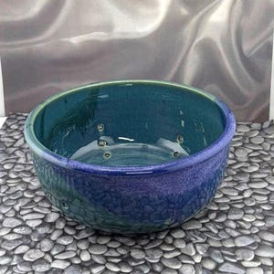 May include: A handcrafted ceramic bowl with a textured exterior and a glossy, teal interior. The bowl has a dark blue rim and a series of small holes around the sides. The bowl is a decorative piece.