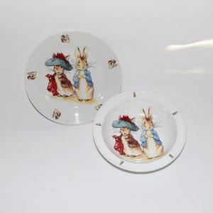 May include: Two white ceramic plates with a Peter Rabbit design. The plates feature illustrations of two rabbits wearing clothes. The larger plate is a dinner plate, and the smaller plate is a bowl. The plates have a white background.