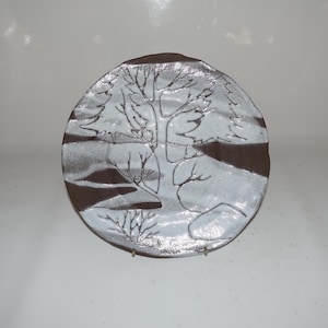 May include: A white ceramic plate with a brown glaze and a tree design. The tree is outlined in white and has a brown trunk. The plate is on a white stand.