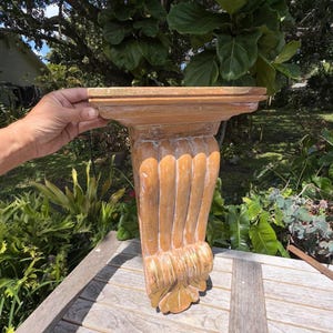 May include: A wooden wall shelf with a decorative, carved design. The shelf is a light brown color with a fluted column and ornate base. The shelf is being held up by a hand.