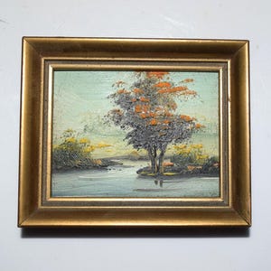 Op de afbeelding: Olieverfschilderij van een landschap met een boom en water, omlijst in een goudkleurige lijst. De boom heeft grijs en oranje blad. Het water weerspiegelt de boom en de lucht. Het schilderij is klein.