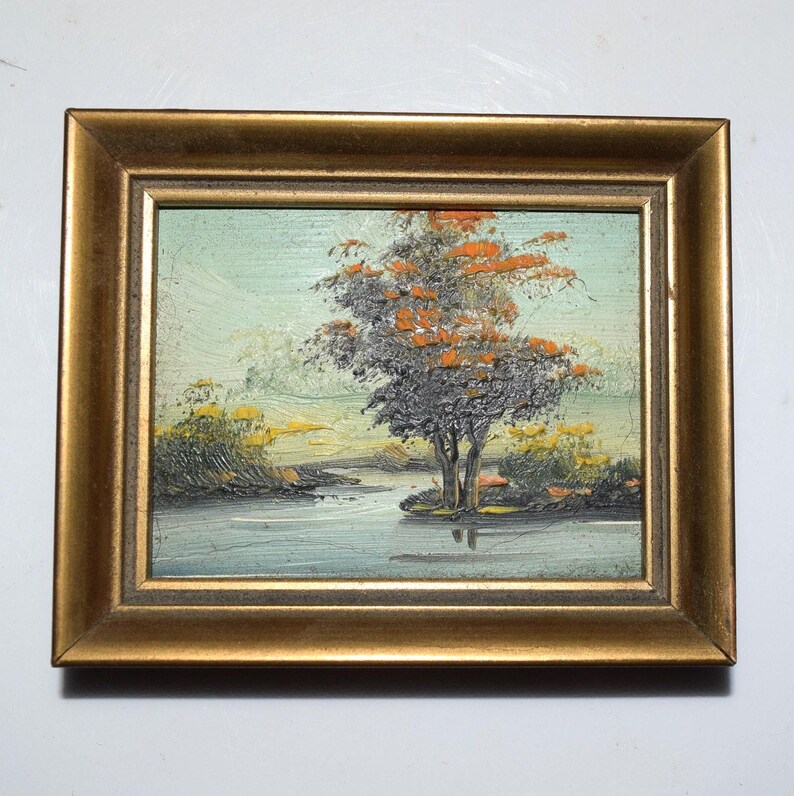 Op de afbeelding: Olieverfschilderij van een landschap met een boom en water, omlijst in een goudkleurige lijst. De boom heeft grijs en oranje blad. Het water weerspiegelt de boom en de lucht. Het schilderij is klein.