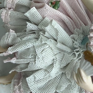 May include: Close-up of a porcelain figurine with a layered design. The figurine features white and pink mesh fabric, with a small green and red accent. The details include ruffled edges and a delicate, textured appearance, suggesting a vintage or antique aesthetic.