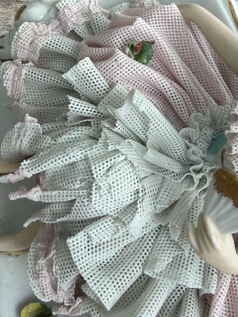 May include: Close-up of a porcelain figurine with a layered design. The figurine features white and pink mesh fabric, with a small green and red accent. The details include ruffled edges and a delicate, textured appearance, suggesting a vintage or antique aesthetic.