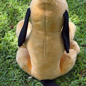 May include: A plush toy dog, viewed from the back, sits on green grass. The dog has a tan body, black ears, and a black tail. The toy is soft and appears well-loved.