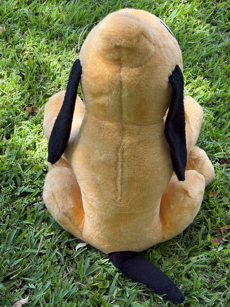 May include: A plush toy dog, viewed from the back, sits on green grass. The dog has a tan body, black ears, and a black tail. The toy is soft and appears well-loved.