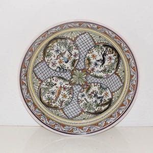 May include: A round ceramic plate featuring a decorative border with orange, blue, and green floral patterns. The center displays four circular medallions containing illustrations of birds and animals, separated by black cross-hatched grid sections.