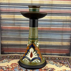 May include: A ceramic candlestick holder featuring a flared base with a multi-colored floral pattern in shades of blue, orange, green, and white. The stem is decorated with vertical green stripes, topped by a dark brown rim and a wide circular drip tray.