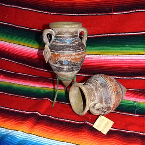 May include: Two antique-style ceramic vases with handles, one upright on a green metal stand, the other lying on its side. The vases have a distressed finish with brown, blue, and white accents. They rest on a colorful striped textile.