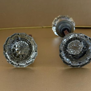May include: Two clear glass door knobs featuring a multi-faceted, starburst-like pattern radiating from the center. Each knob has a circular, scalloped edge and is attached to a dark metal base with a square spindle extending from the back.
