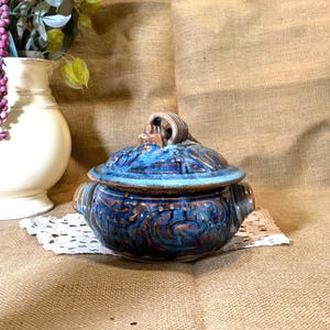 May include: A round ceramic lidded casserole dish featuring a glossy glaze in shades of deep blue and earthy brown. The surface displays swirling patterns and raised dot textures. It has two side handles and a central handle on the lid.