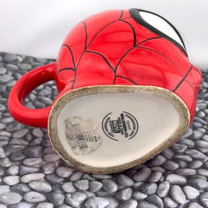 May include: A red ceramic mug featuring a Spider-Man design. The mug has a red exterior with black web lines and a white and black eye design. The handle is red. The bottom of the mug has text that reads "Dishwasher Safe" and "Microwave Safe".