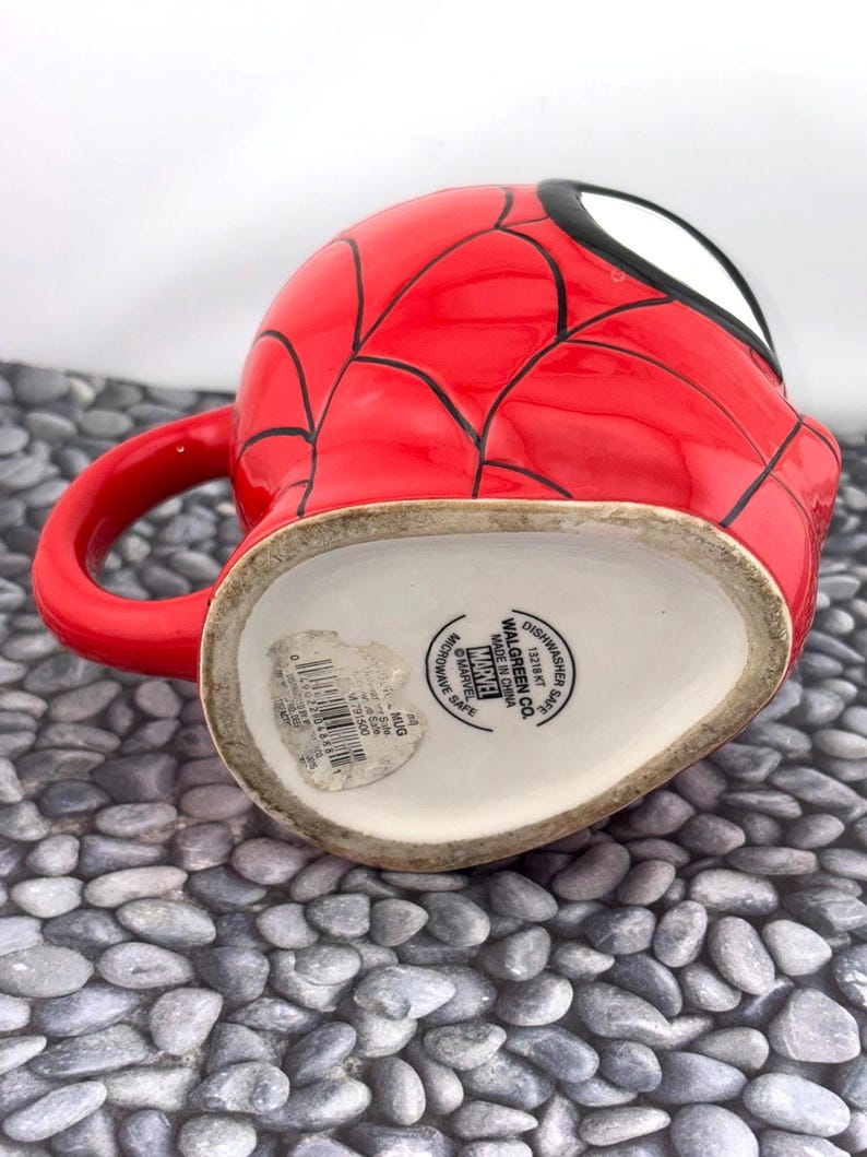 May include: A red ceramic mug featuring a Spider-Man design. The mug has a red exterior with black web lines and a white and black eye design. The handle is red. The bottom of the mug has text that reads "Dishwasher Safe" and "Microwave Safe".