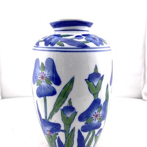 May include: White ceramic vase with a blue floral design. The vase features blue flowers with pink centers and green leaves. The rim and top of the vase are also decorated with blue floral patterns. A decorative piece for the home.