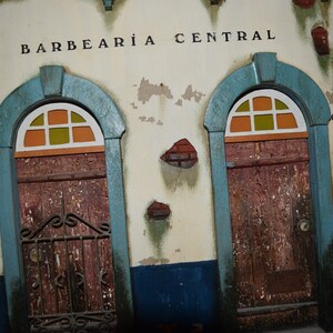 May include: A weathered building facade with two arched doorways, each with a stained glass window and a wooden door. The building is painted white with blue trim and has the words "BARBEAR&Iacute;A CENTRAL" written above the doorways.