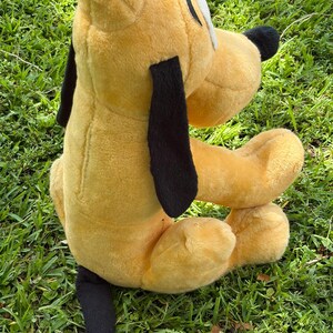 May include: A plush toy of Pluto, a yellow dog with black ears and tail. The toy has a white patch on its face and a black nose. The toy is sitting on green grass.