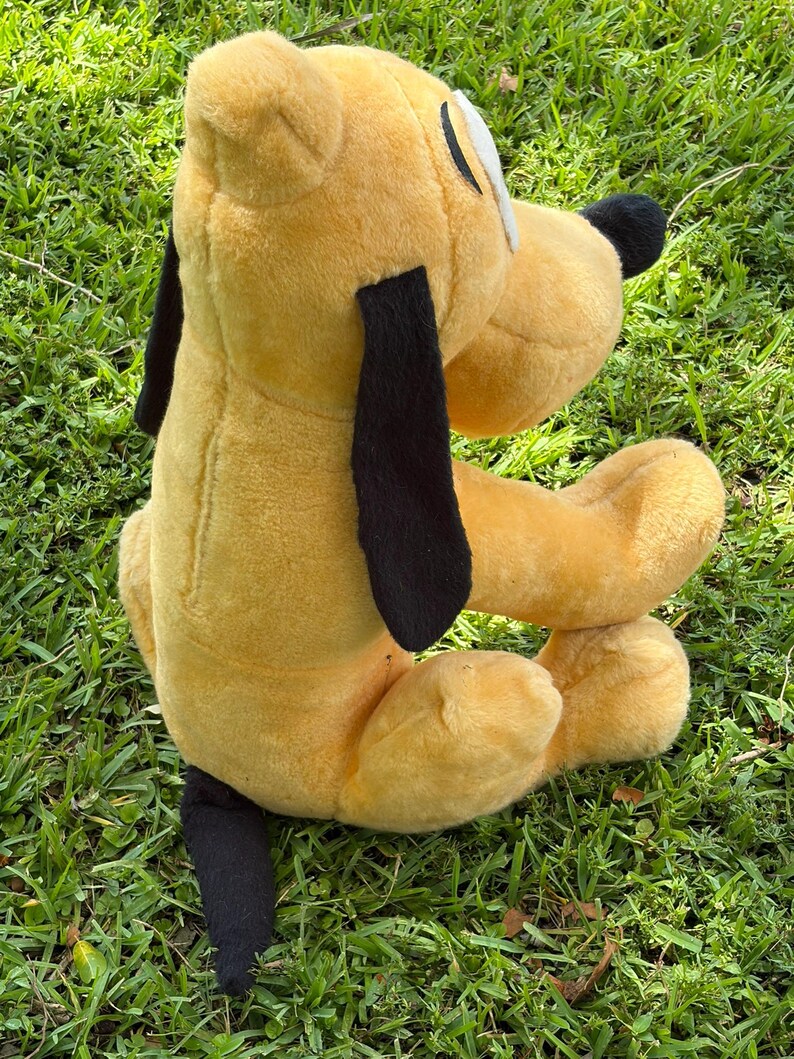 May include: A plush toy of Pluto, a yellow dog with black ears and tail. The toy has a white patch on its face and a black nose. The toy is sitting on green grass.