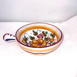 May include: A small, round ceramic bowl with a handle, featuring a floral design. The bowl has a white interior with a yellow and orange floral pattern. The exterior has horizontal stripes in orange and white. The handle is white with blue and red accents.
