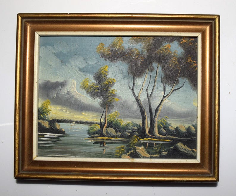 Op de afbeelding: Olieverfschilderij van een landschap met een rivier, bomen en bergen. Het schilderij zit in een goudkleurige lijst. De lucht is een mix van blauw en grijs, en de bomen hebben groene en bruine bladeren.