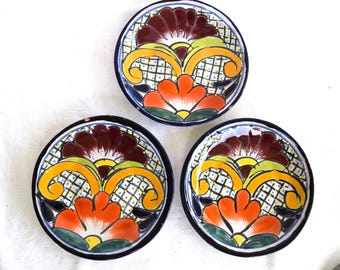 May include: Three small, round, hand-painted ceramic plates. Each plate features a central floral design in orange, yellow, green, and burgundy, set against a white background with a grid pattern. The plates have a dark blue border.