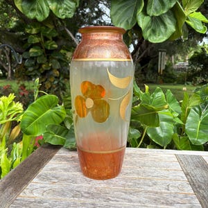 May include: A glass vase featuring a frosted body with a wide, rounded silhouette. The neck and base are finished in a deep amber hue with a textured appearance. A gold-toned floral motif with a single large flower and leaf adorns the center.