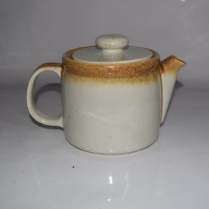 May include: A white ceramic teapot with a brown speckled glaze and a brown rim. The teapot has a lid and a handle.