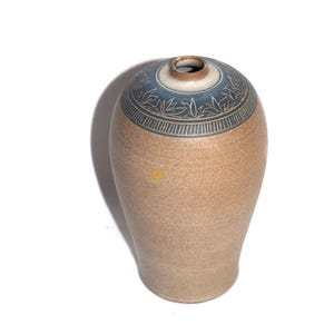 May include: A stoneware vase with a speckled tan body and a narrow neck. The upper shoulder features a dark blue band decorated with a carved leaf pattern and vertical lines. The rim is finished in a smooth, unglazed brown tone.