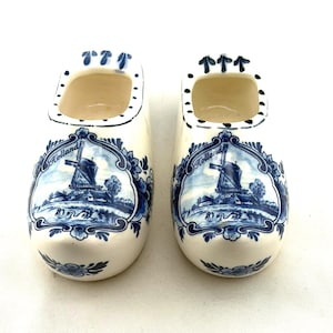 May include: Pair of ceramic Dutch clogs, painted white with blue floral and windmill designs. The clogs have the word "Holland" in a decorative banner. The clogs are a traditional souvenir item.
