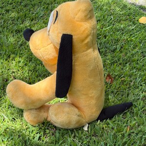May include: A plush toy dog in a seated position, featuring a yellow body, black ears, and a black tail. The dog has a white eye and is resting on green grass.