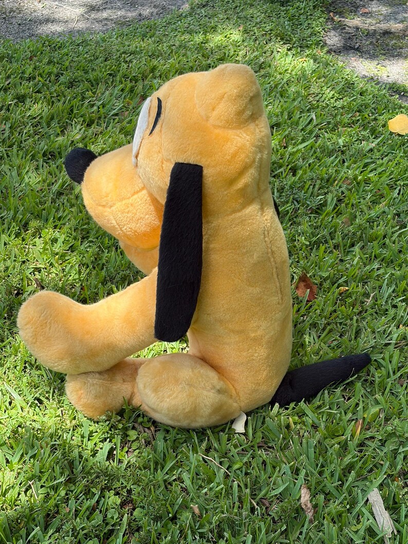 May include: A plush toy dog in a seated position, featuring a yellow body, black ears, and a black tail. The dog has a white eye and is resting on green grass.