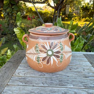May include: A round, terracotta-colored ceramic pot with a lid. The pot features a floral design in white, green, and brown. It has two handles and a knob on the lid. The pot is sitting on a wooden table.
